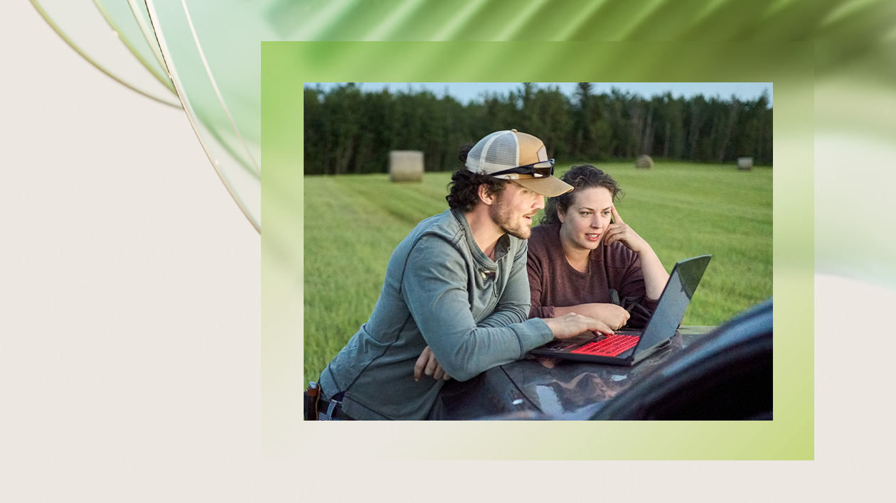 Two people looking at a laptop on the hood of a car parked in a hayfield