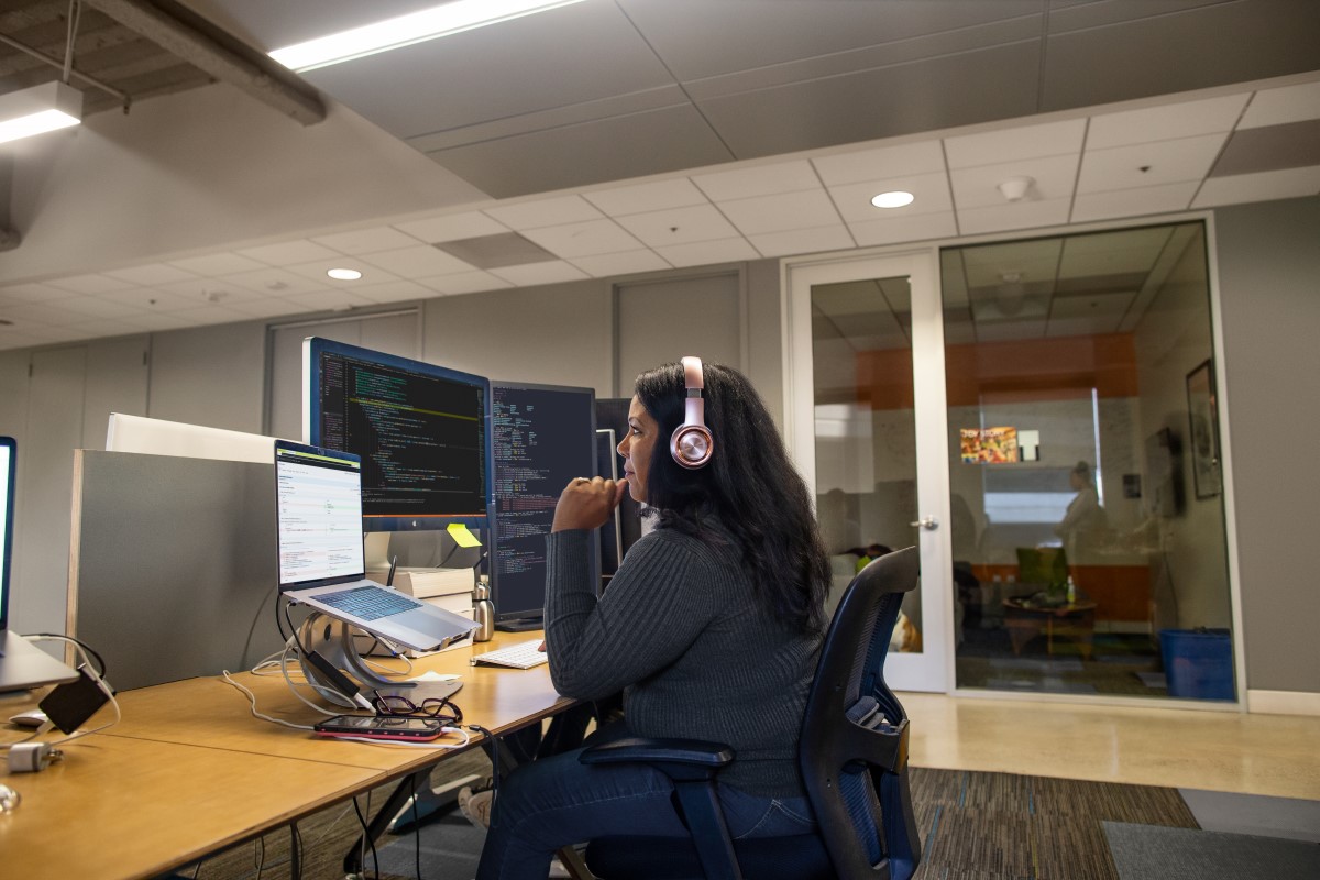 Real people, real offices. Black female developer wearing headphones, coding at her desk