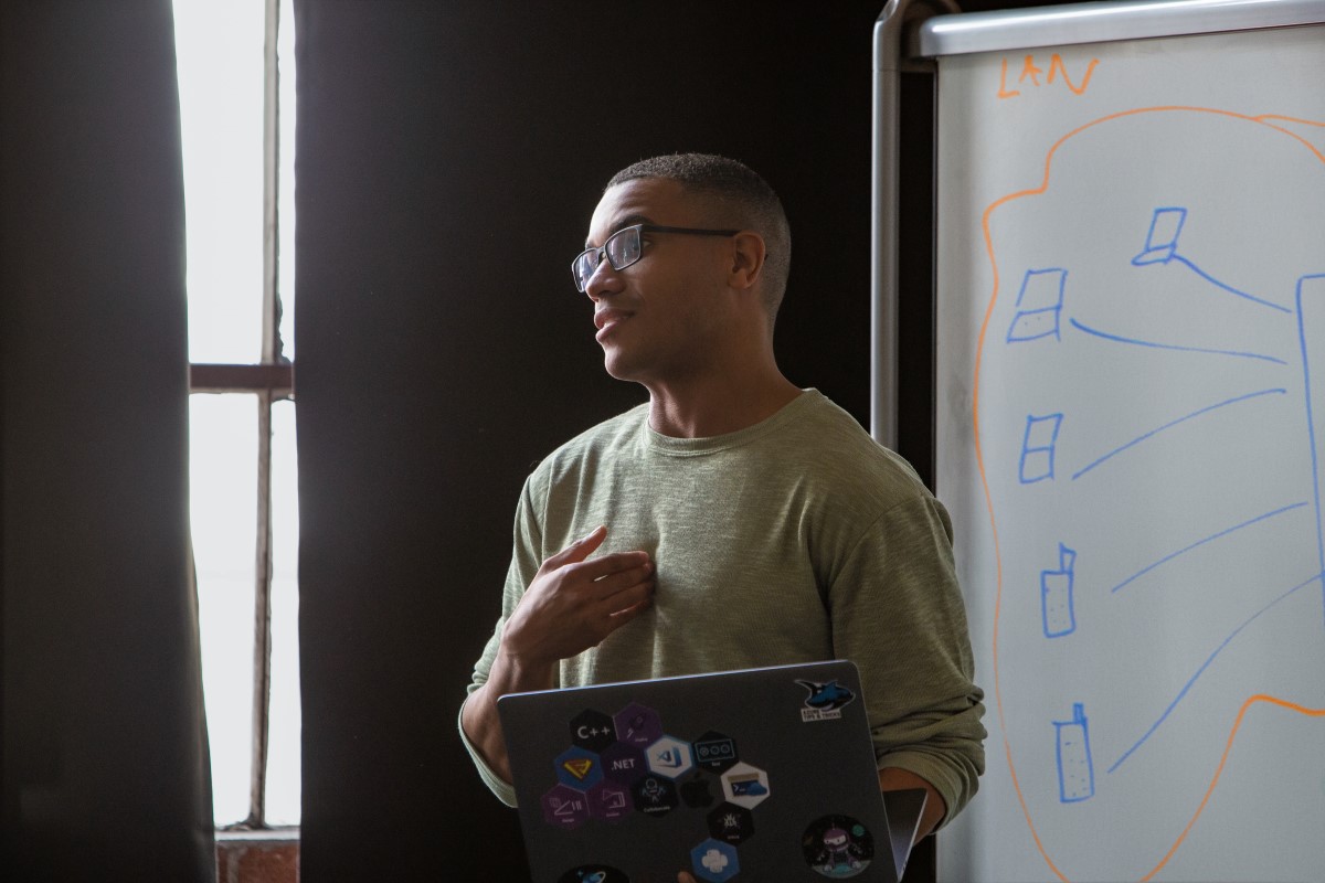 Real people, real offices. Male developer speaking in front of a white board during team stand up meeting, holding a Surface laptop personalized with stickers. Code, develop, developer, engineer, Visual Studio, Azure