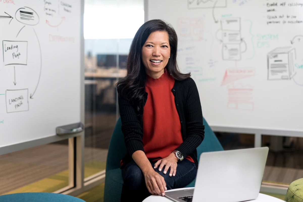 Portrait of female executive in red shirt, smiling and facing camera. She is in an office conference room setting, using an open laptop (screen not shown). Two whiteboards are partially visible in the background, with diagrams and flowcharts written on them