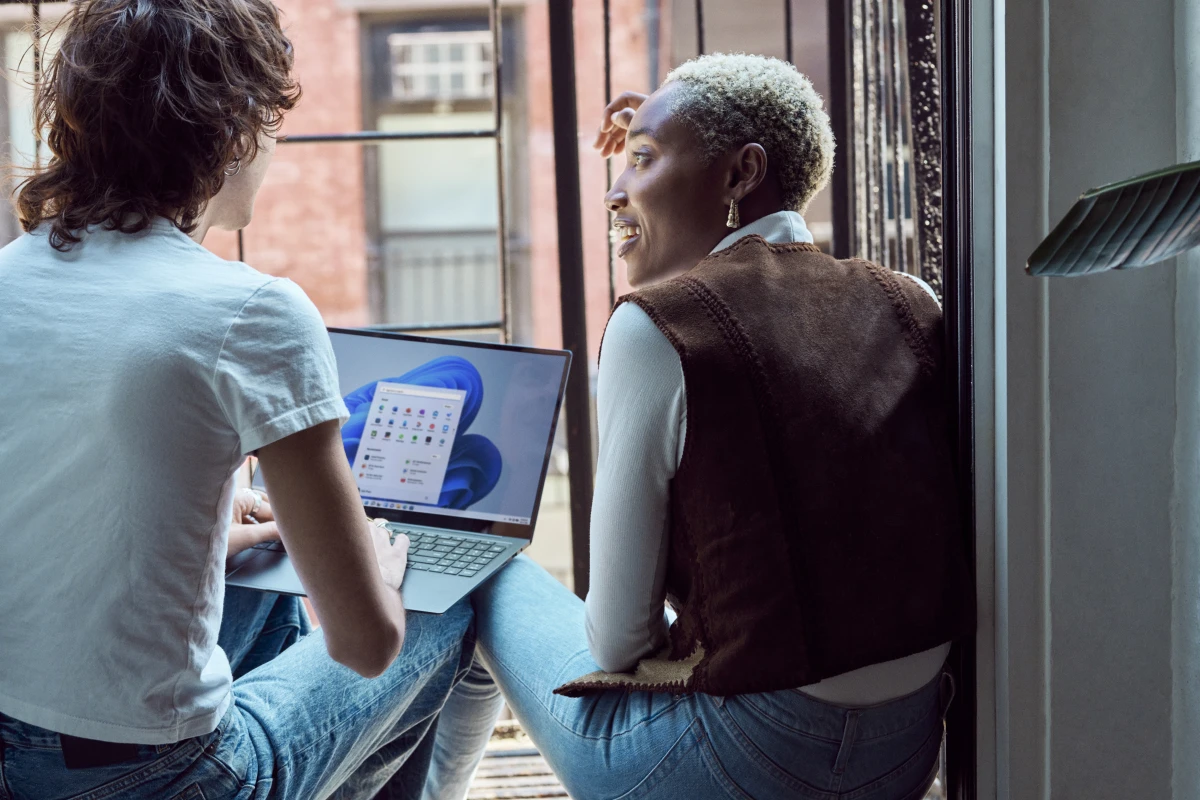 Two Gen Z friends sitting on fire escape balcony with backs to camera laptop open with Windows 11 start screen and start menu