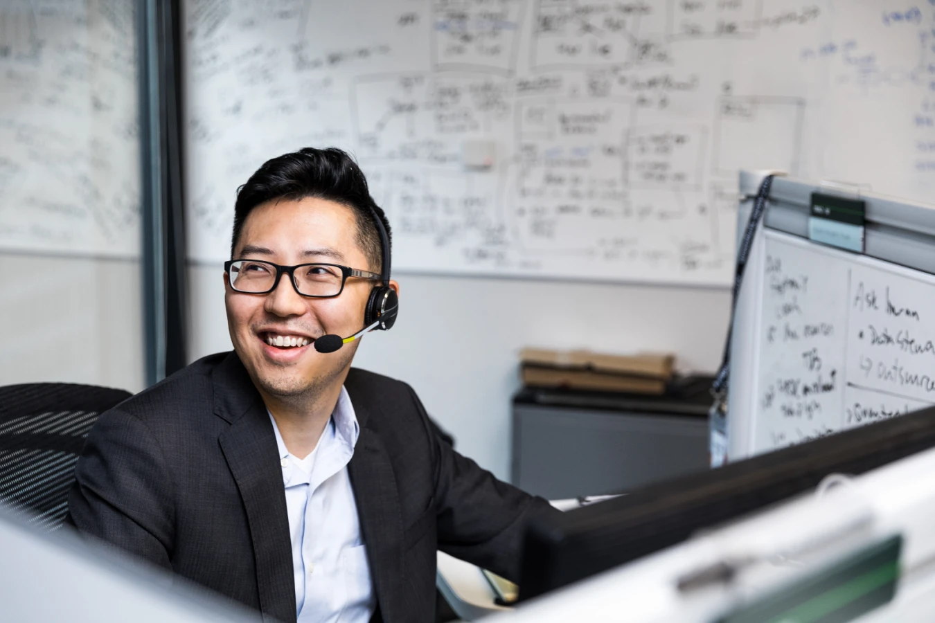 Male worker sitting at cubicle desk in financial office, wearing glasses and phone headset. He is smiling and looking up to his right. A large whiteboard covered in black written text is on wall behind him. A smaller whiteboard also partially visible on his desk‘s cubicle wall