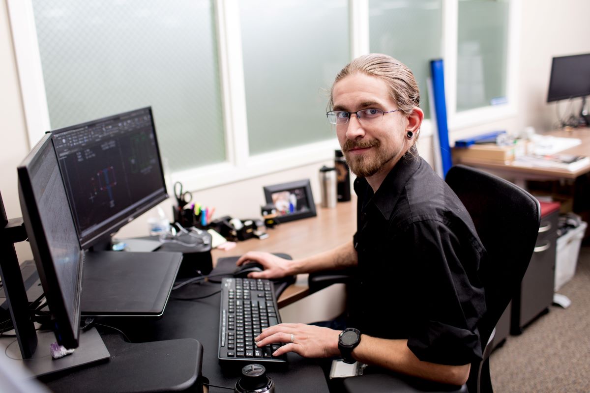 Portrait of male worker in manufacturing office sitting at desk in front of two desktop monitors. He is looking at camera, with one hand on the keyboard and one hand on the mouse. Screens show basic diagrams