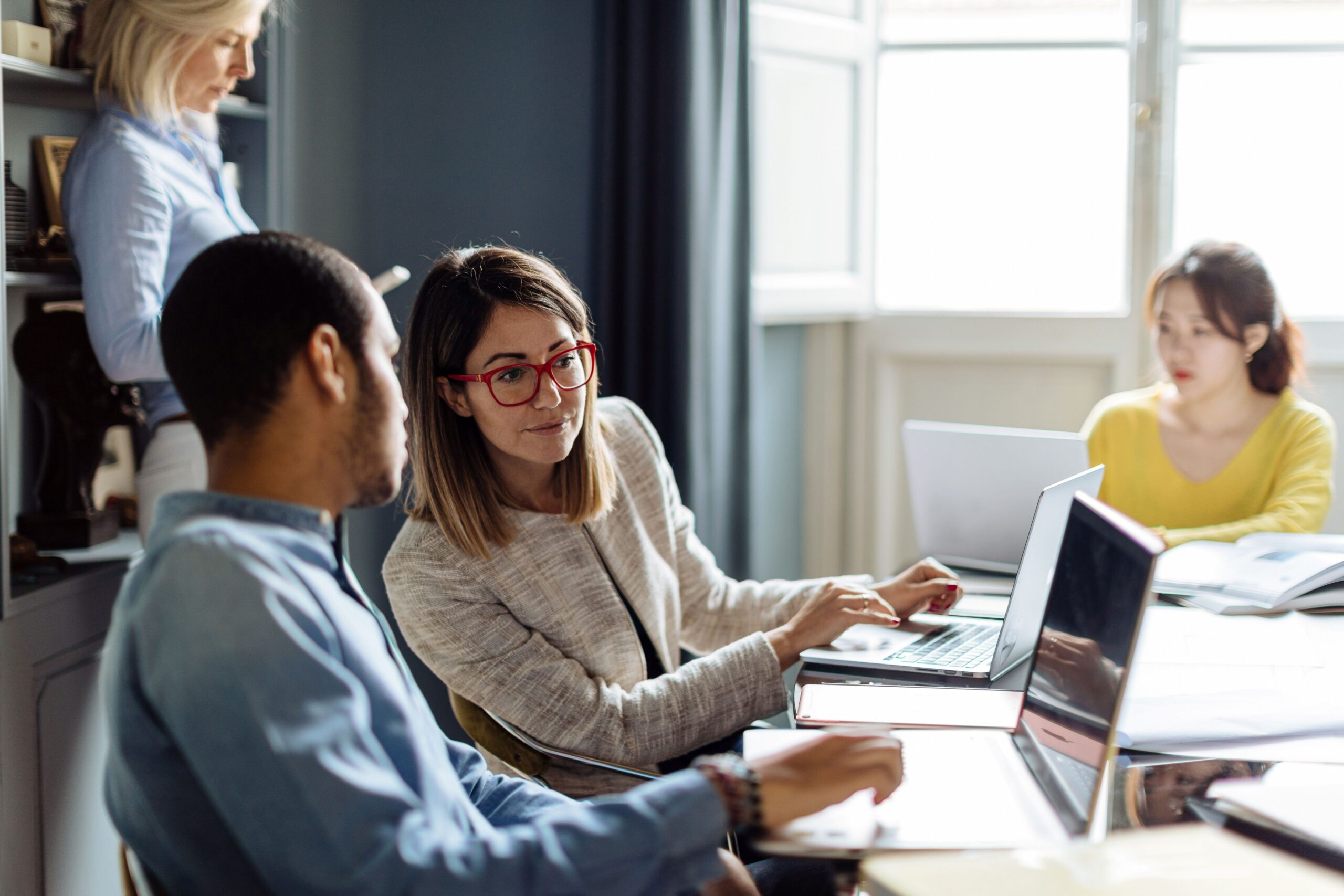 Small group of professionals collaborating at a table with laptops and notebooks, discussing ideas in a bright office meeting space