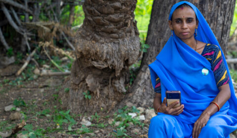 Lady in blue sari, holding a smartphone, looking at the camera. Seated outside in front of a couple of trees.