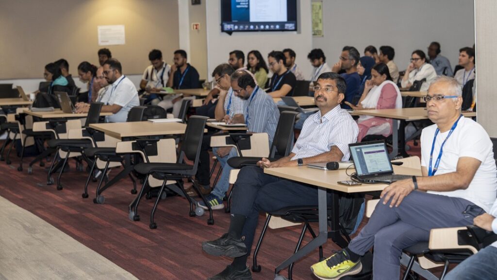 Photograph of a conference room with an audience seated at tables facing a stage. The setting includes wall-mounted screens displaying content, indicating an ongoing presentation or lecture.