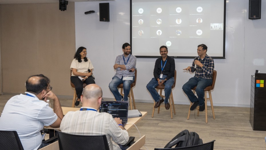 Photograph of a panel discussion in a conference room with four speakers seated on high chairs at the front and an audience seated at tables facing them. The setting includes a projector screen displaying a presentation, and attendees engaged in listening and note-taking.