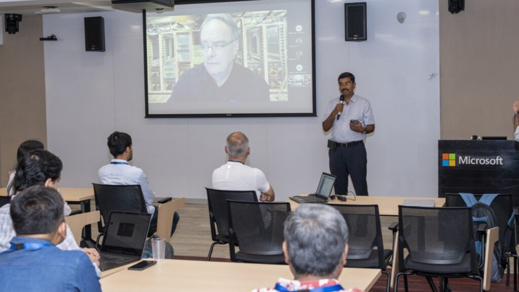 Photograph of Venkat Padmanabhan giving a presentation in a conference room with a Microsoft logo on a podium in the foreground. The presenter holds a microphone and gestures toward a projected slide displaying remote attendees.