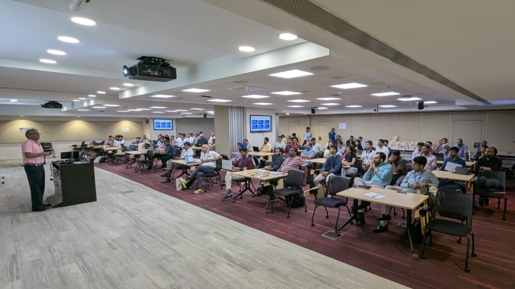 Photograph of a conference room with an audience seated at tables facing a speaker standing near a podium. The setting includes ceiling lights, wall-mounted screens displaying content, indicating an ongoing presentation or lecture.