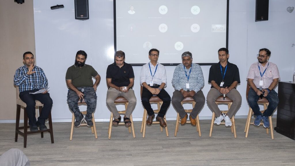 Photograph of a panel discussion featuring six men seated on high stools in a conference room, with a projection screen displaying a virtual meeting interface behind them. The setting suggests a hybrid event combining in-person and remote participation, with panelists dressed in business casual attire and wearing conference badges.