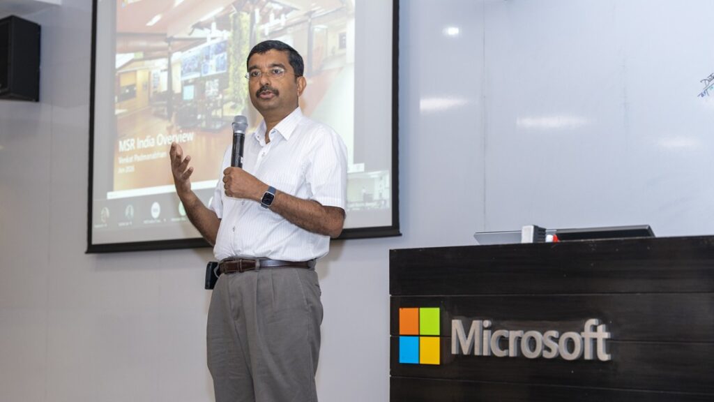 Photograph of Venkat Padmanabhan giving a presentation in a conference room with a Microsoft logo on a podium in the foreground. The presenter holds a microphone and gestures toward a projected slide displaying 