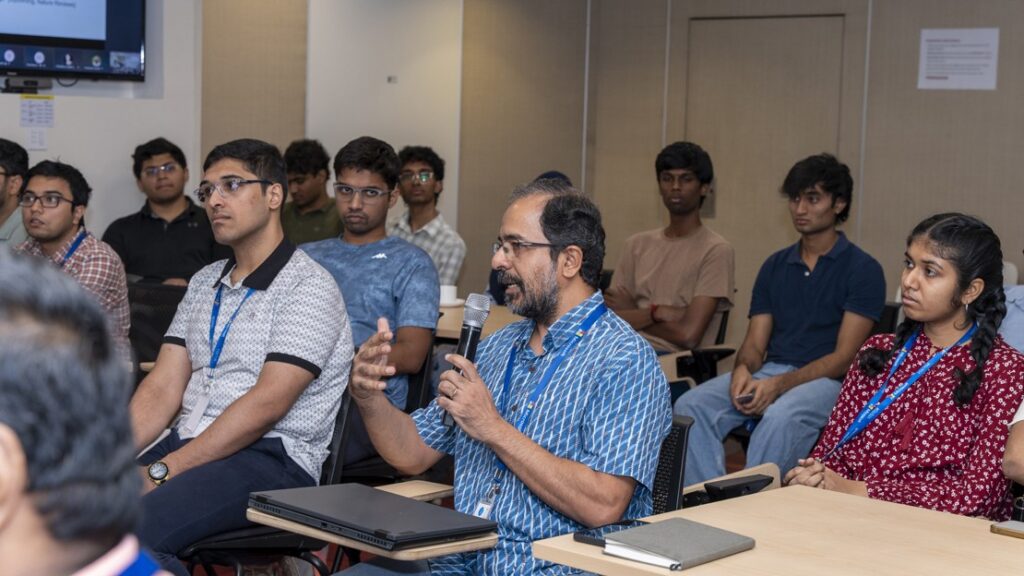 Photograph of a group of people seated in a classroom or seminar setting, with one man in a blue patterned shirt holding a microphone and speaking. Attendees wear casual clothing and lanyards, with notebooks and laptops on desks, indicating an interactive discussion or presentation.