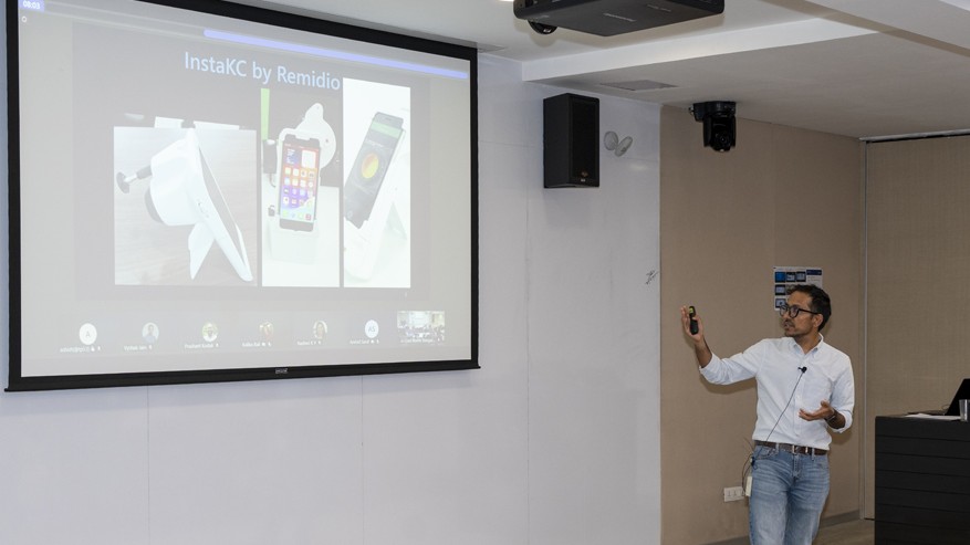 Photograph of Mohit Jain giving a presentation in a conference room, pointing at a large screen displaying a slide titled 