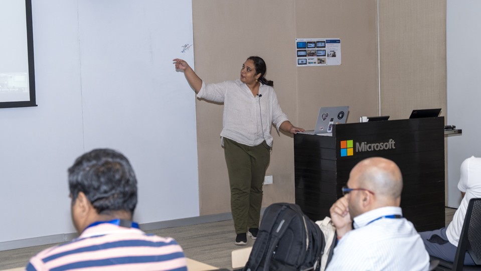 Photograph of Sunayana Sitaram giving a presentation in a Microsoft-branded conference room, pointing towards a screen while attendees listen and take notes. The setting includes a podium with a laptop, several seated participants, and a neutral-colored background with minimal decor.