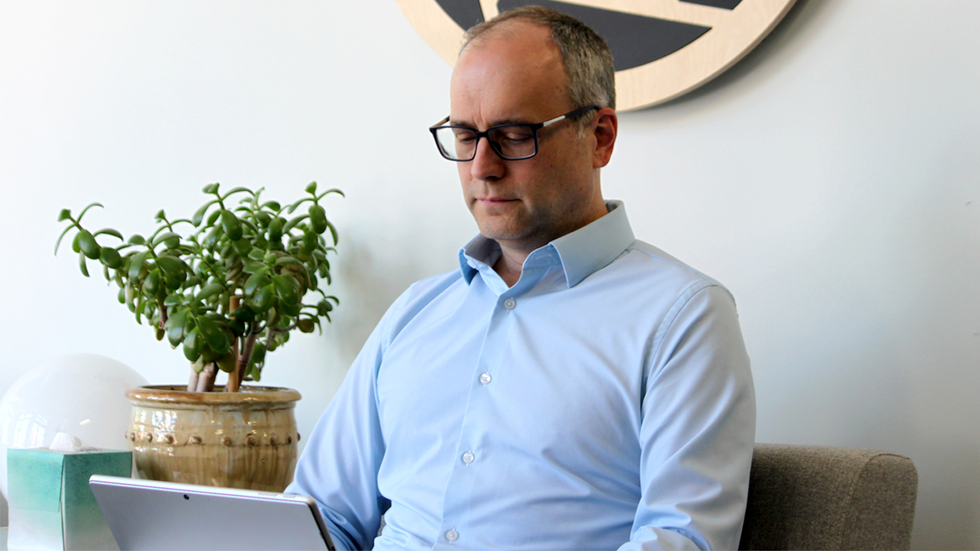 Kurt DeMaagd seated indoors using a tablet, with a potted plant and desk items nearby, suggesting a workspace environment focused on reviewing or interacting with digital content.