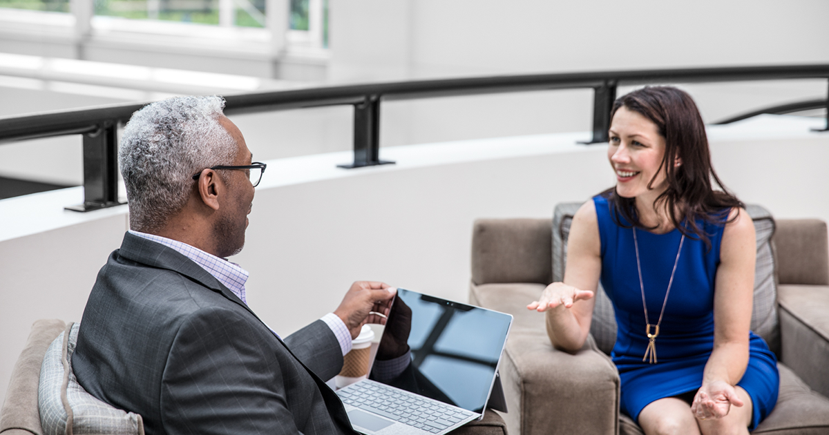 Image of two office workers talking. One has a laptop open