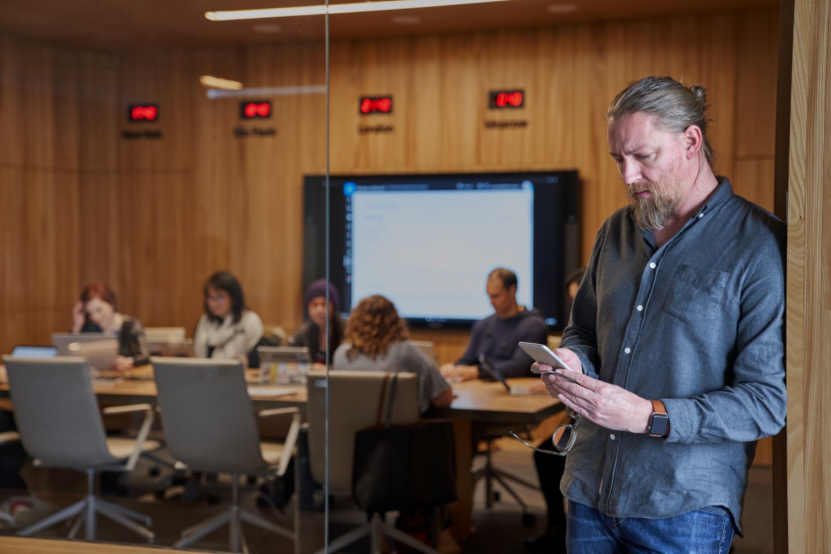 Bearded man looking at his phone with background of a blurry conference room of people working