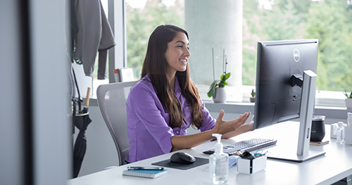 An image of a female employee talking on a video call with hand sanitizer on her desk, socially distancing from others