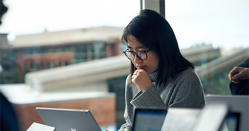 An image of a woman with glasses sitting at a community table in common area reading on a Surface laptop