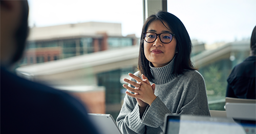An image of a woman with glasses sitting at a community table in common area looking up from behind a Surface laptop with her hands clasped together