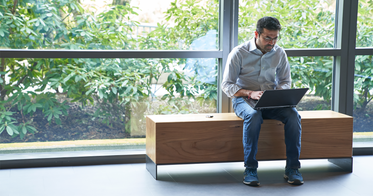 An image of a man sitting on an indoor bench in front of windows working on a laptop with a security key fob or USB drive next to him on the bench