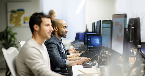 An image of two male coworkers sitting next to each other at along row of office desks, using their own desktop computers