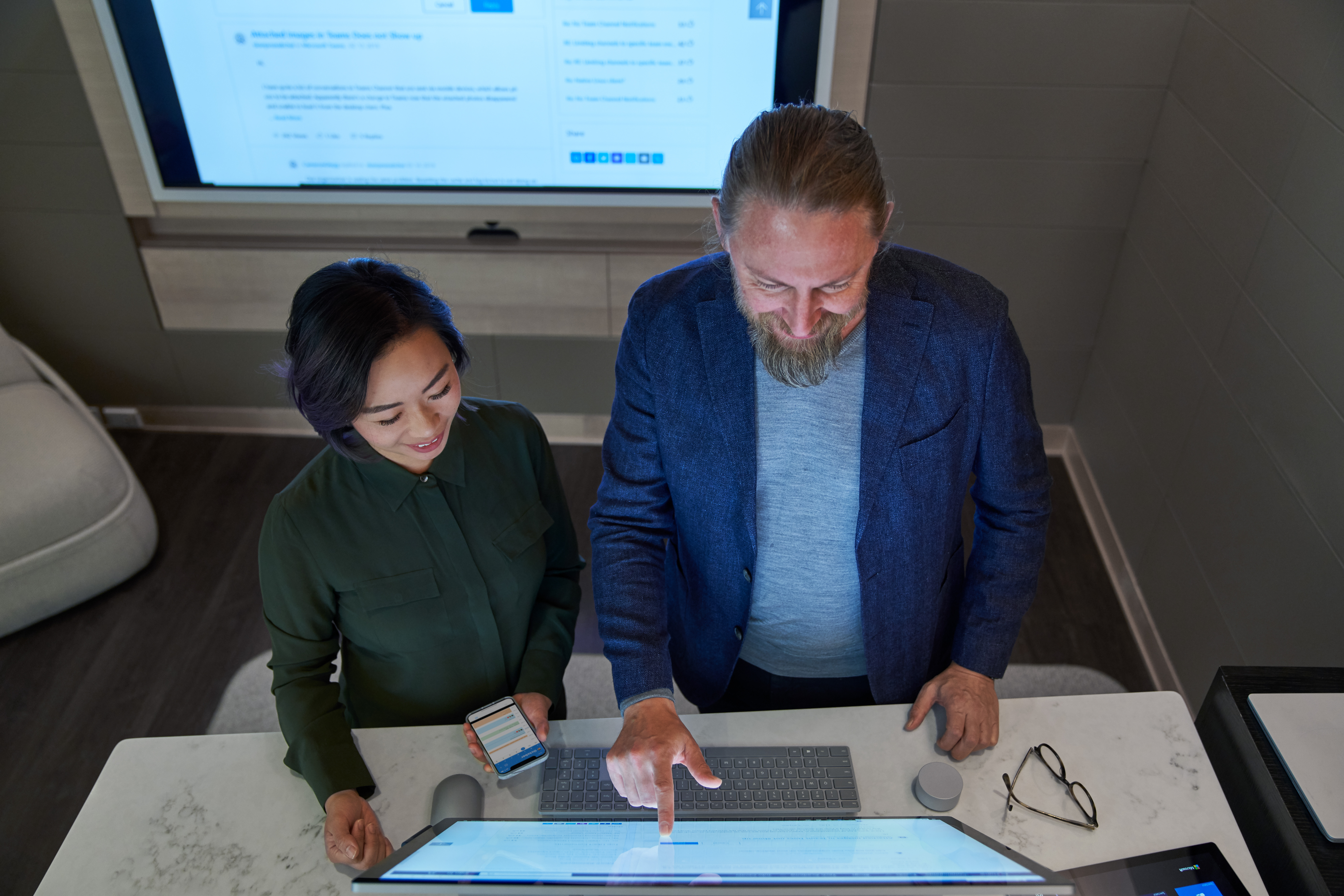 Top down view of a man and woman in a dim office collaborating or working on a Microsoft Surface Studio. She is holding a phone