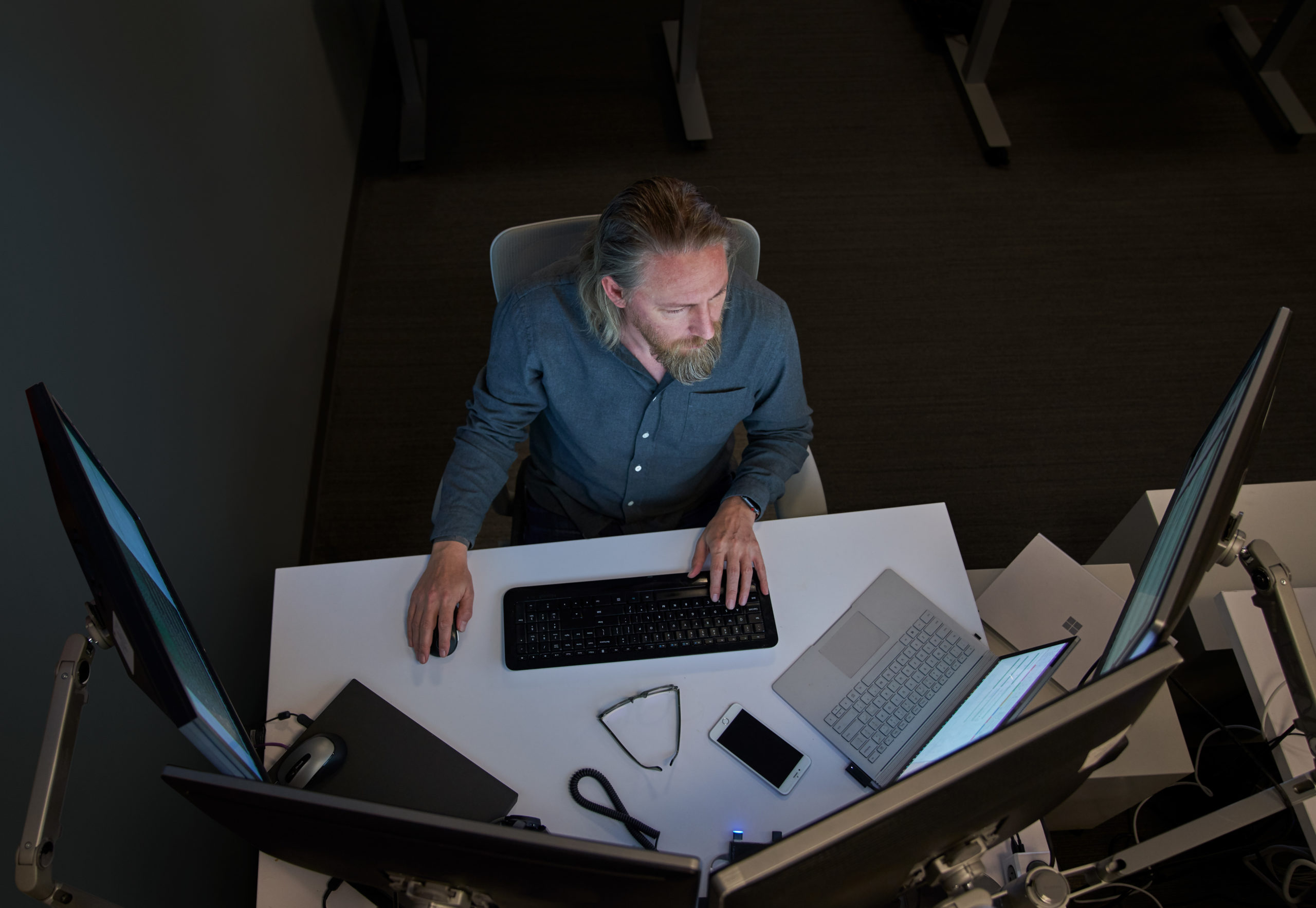 Top-down view of a bearded man in a gray/blue shirt seated at a desk working on a Surface laptop connected to three large monitors