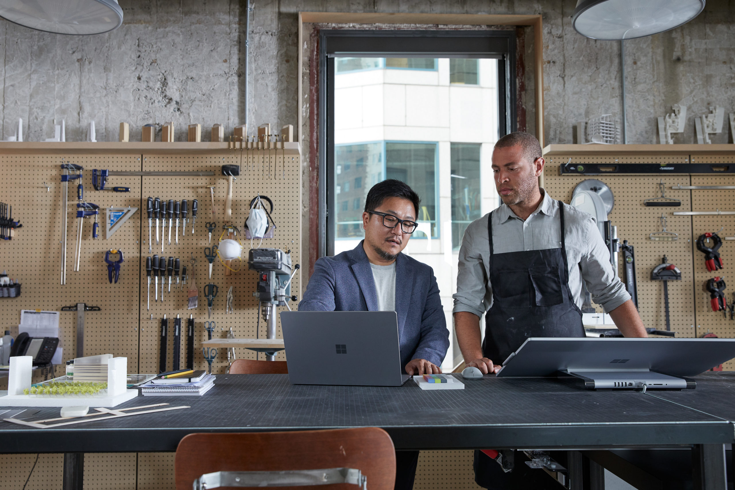 Two adult male collaborating in architecture modeling studio looking together on a platinum Microsoft Surface Laptop 3 and a platinum Microsoft Surface Studio 2