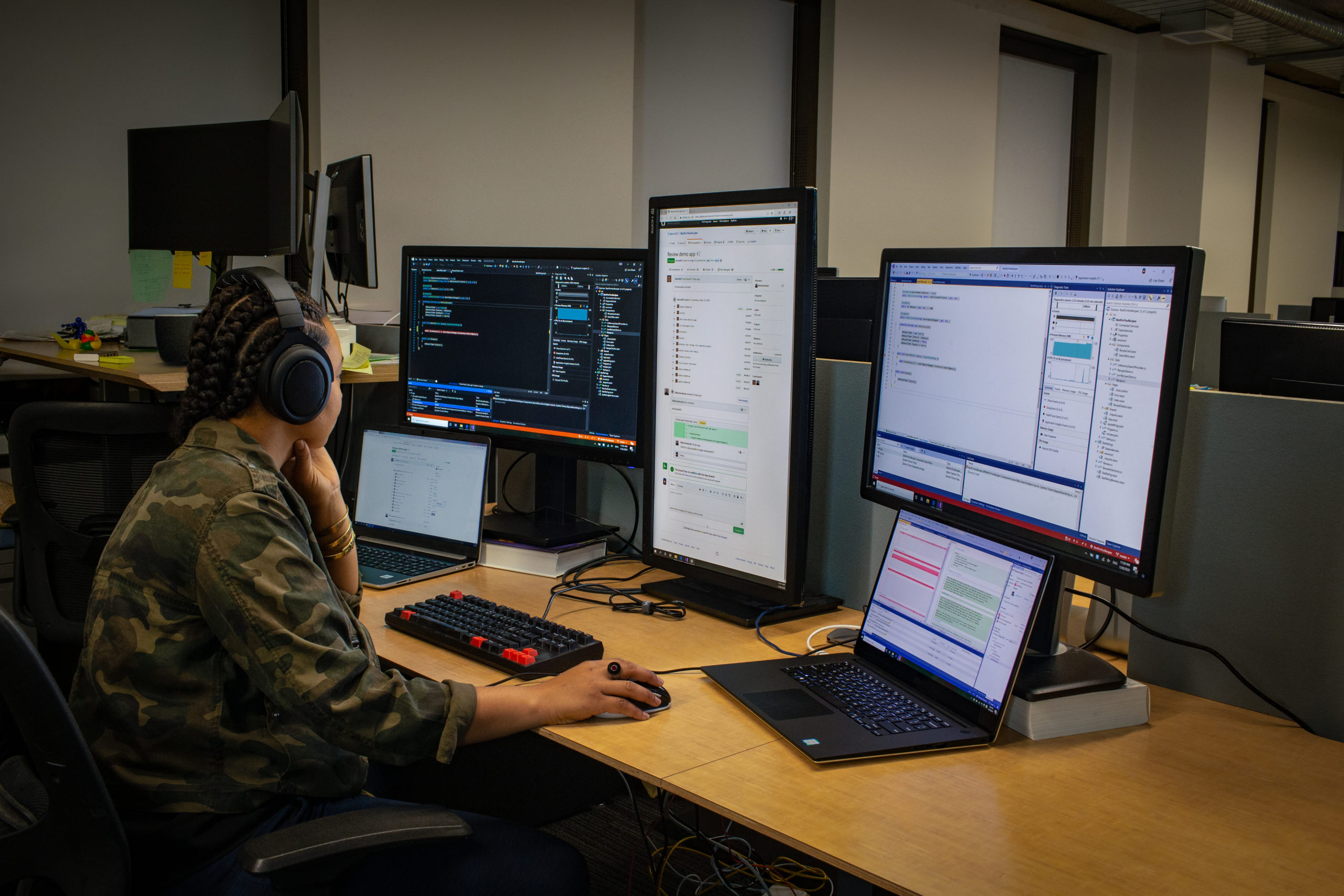 lack female developer wearing headphones; coding at her PC workspace in an enterprise office, using Visual Studio on a multi-monitor set up
