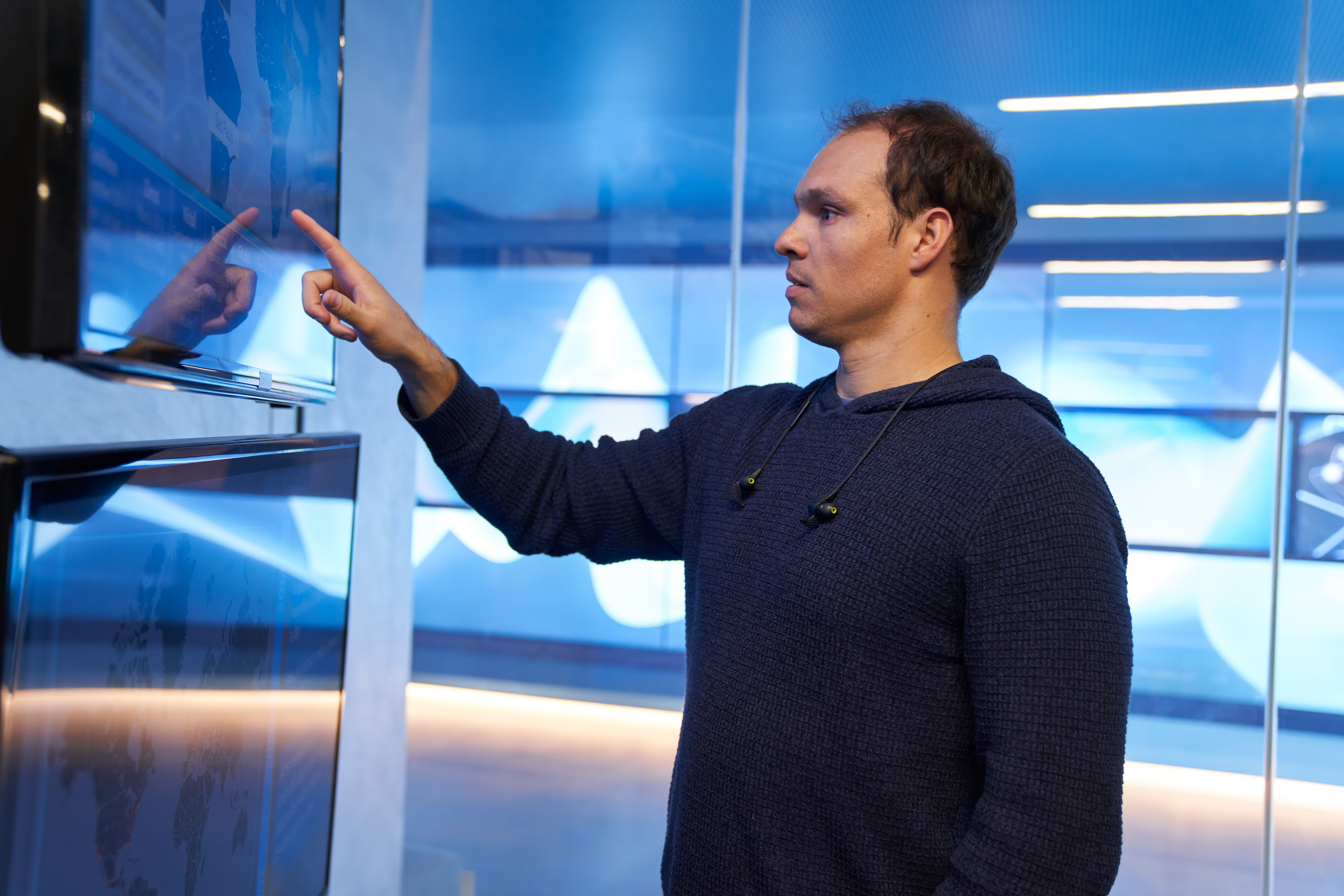 Man in a hooded sweater/sweatshirt inside a secure room, pointing at a geographic area displayed on a large monitor