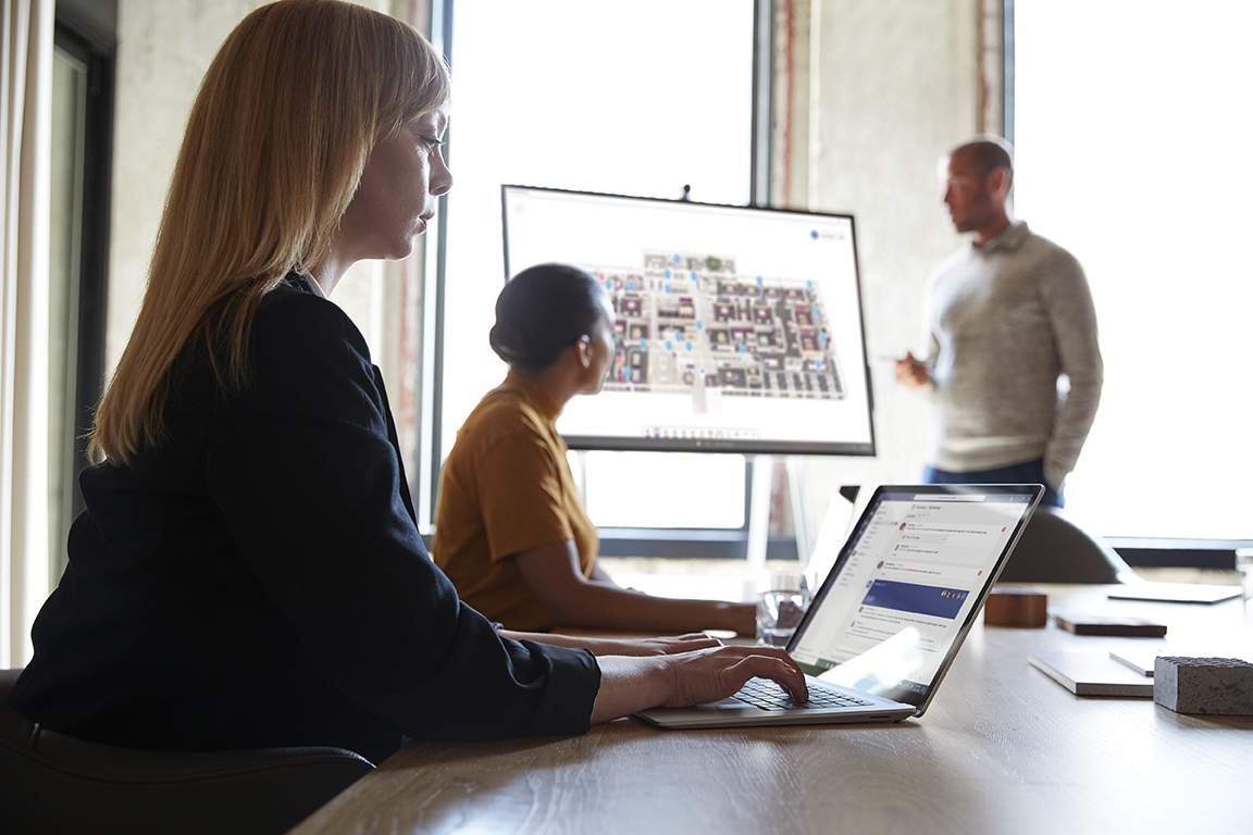 Three adults are collaborating in a conference room. One male is standing next to a Microsoft Surface Hub 50&rdquo; with Microsoft Whiteboard screen shown