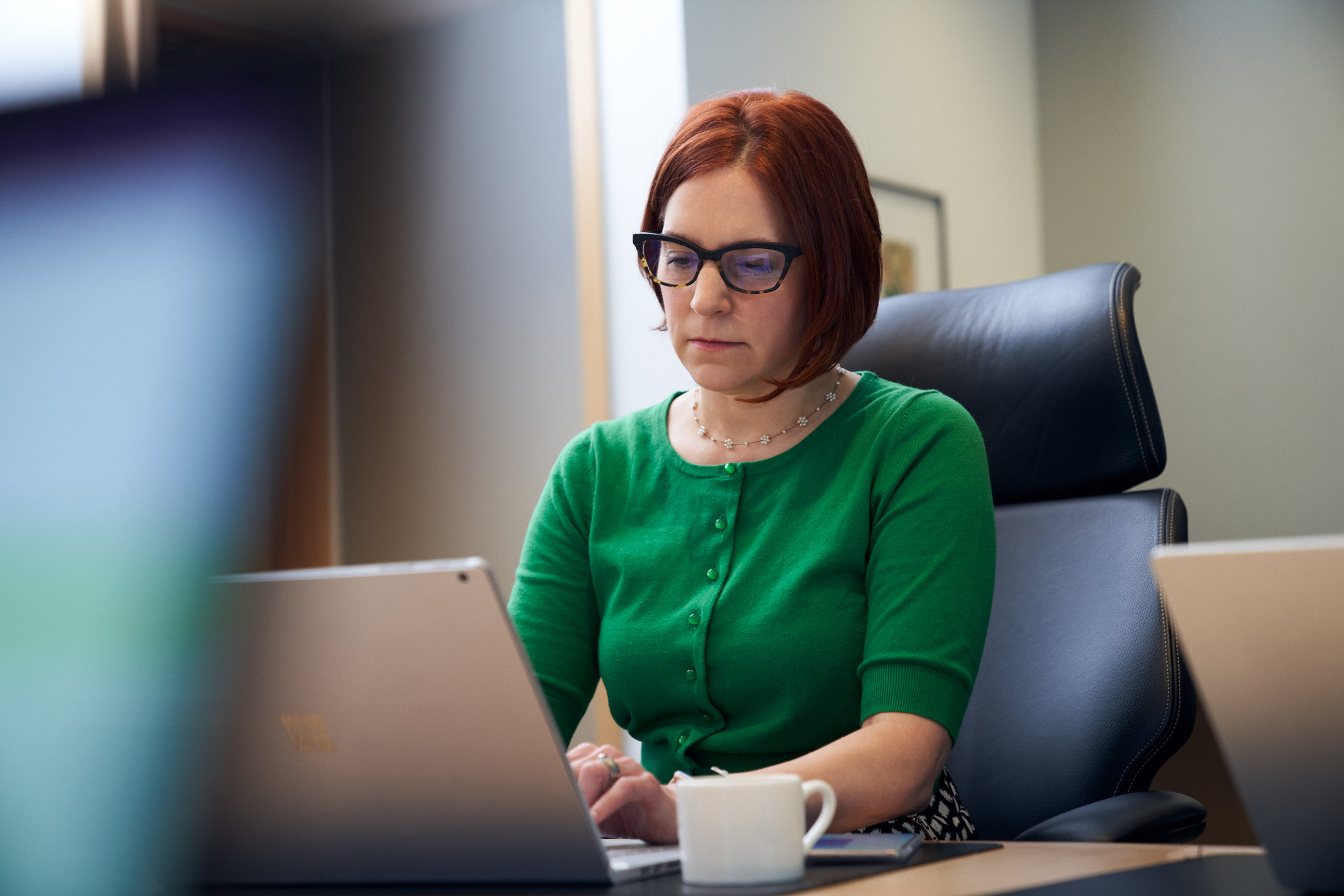 A red-haired woman with a green shirt and glasses, sitting inside an office working on her laptop