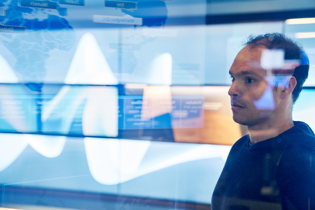 Man in hooded sweater/sweatshirt inside a secure room who is looking at data and a geographic area displayed on a large monitor which is behind glass walls with reflections