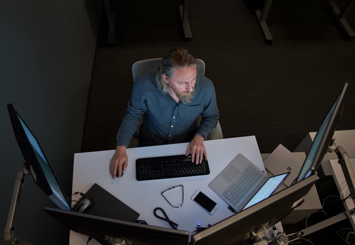Top-down view of a bearded man in a gray/blue shirt seated at a desk working on a Surface laptop connected to three large monitors. Desktop has phone, glasses, mouse and other laptops on it