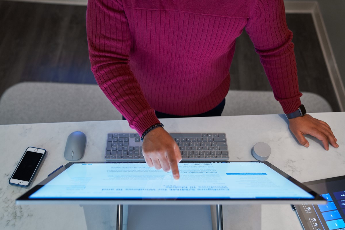 Top down view of a man wearing a dark red shirt working on a Microsoft Surface Studio with a phone next to him on the desktop along with a mouse, keyboard, and Surface Dial