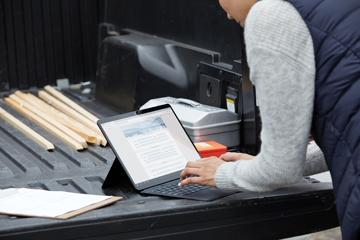 Adult female standing at the back of a pickup truck using a black Microsoft Surface Pro X in laptop mode with Microsoft Edge screen shown