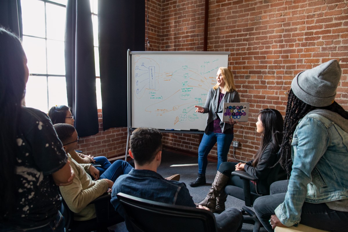 Female developer speaking in front of a white board during team stand up meeting, holding a Surface laptop personalized with stickers
