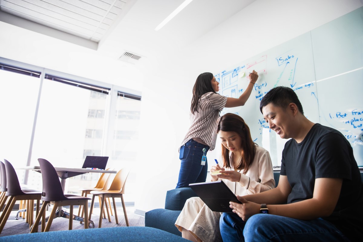 A group of coworkers collaborate on project in conference room of U.S. office. Woman writes on white board while two other coworkers work on a laptop together