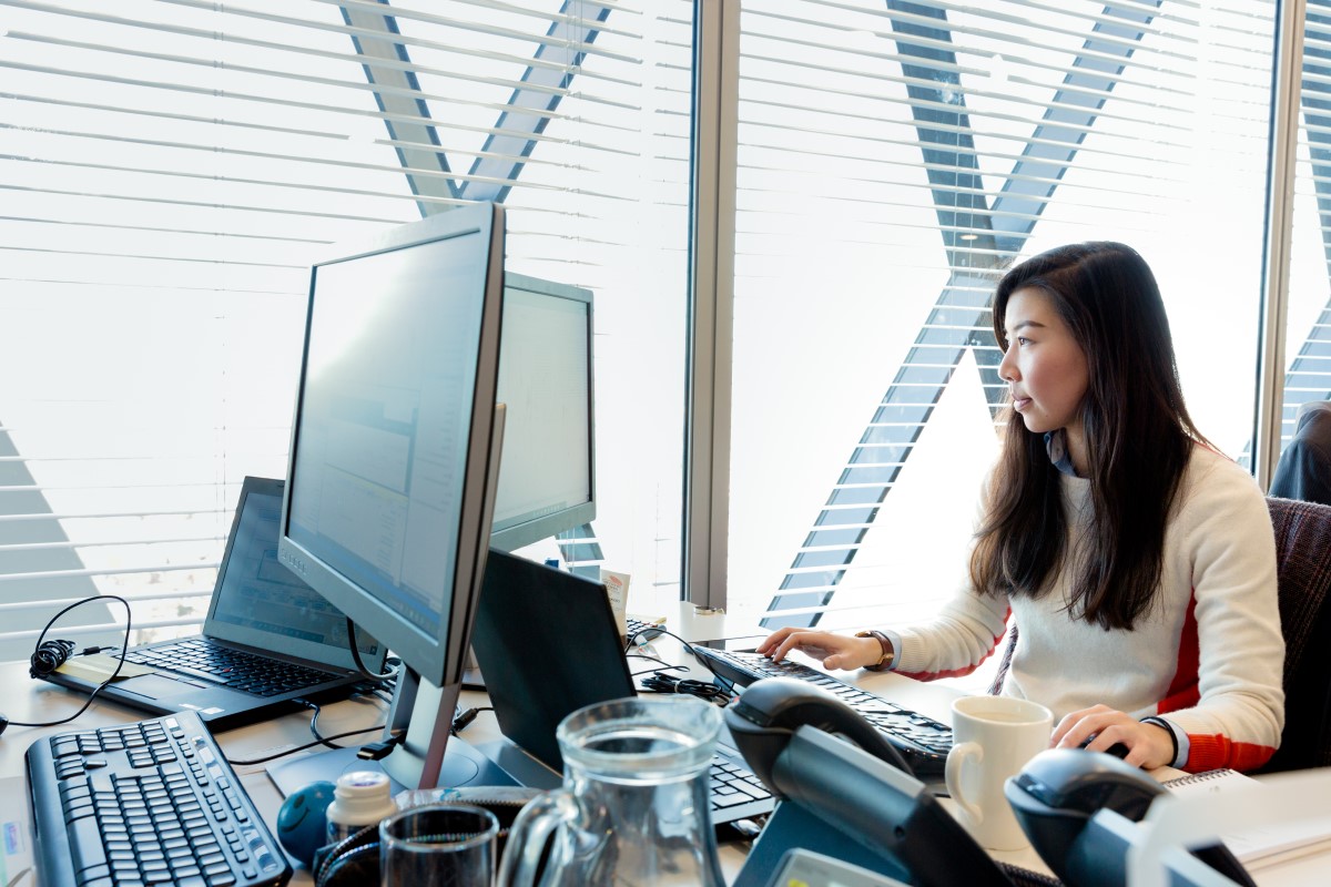 Female office worker at workstation near window, looking at desktop monitor (screen not shown). Open laptop also on desk (screen not shown)