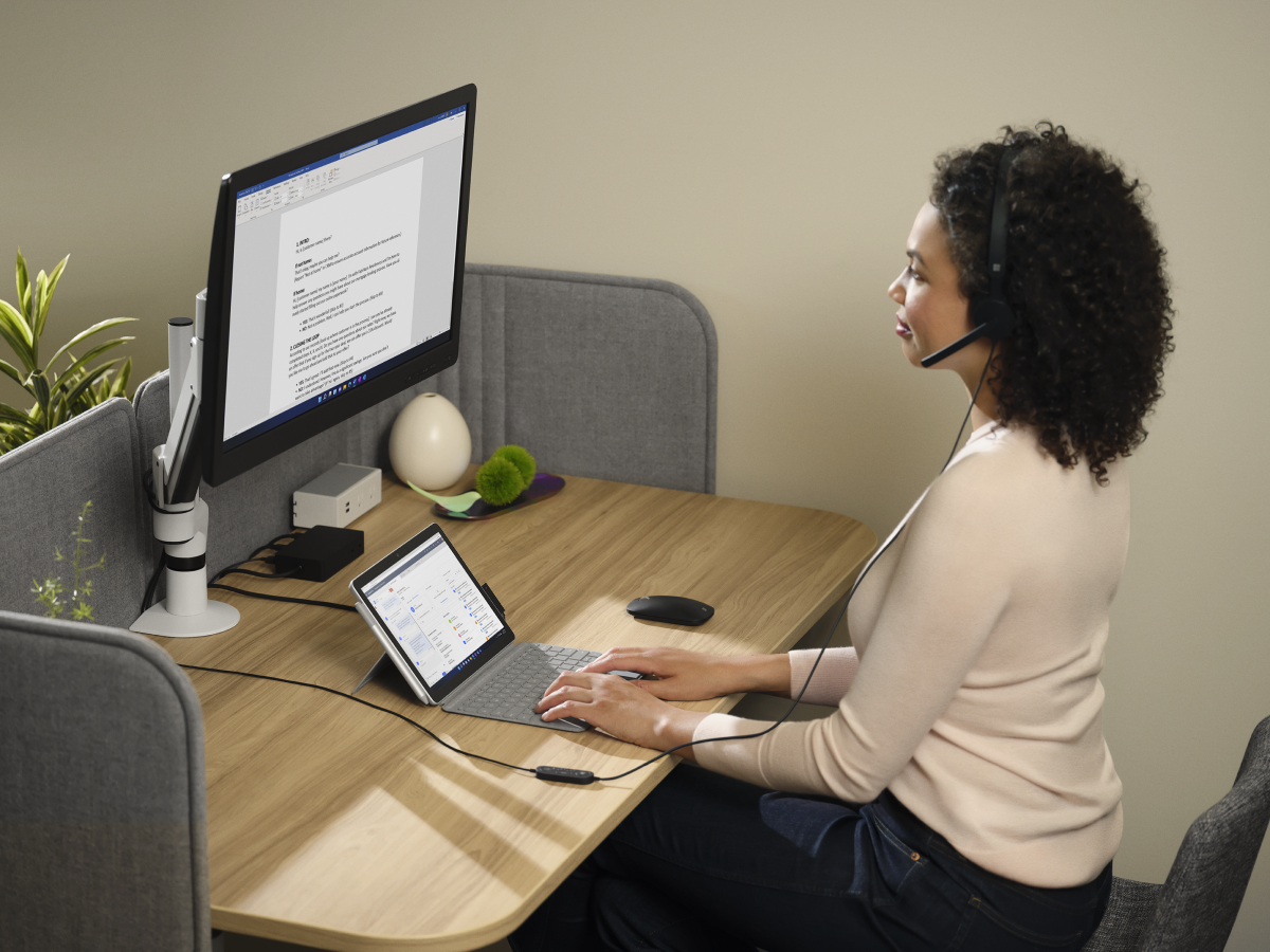 Adult female wearing Microsoft Modern USB-C Headset sitting at a desk while working on a Surface Go 3 with Surface Go Typecover in laptop mode with Microsoft Edge displayed. Microsoft Word is being presented on the large monitor