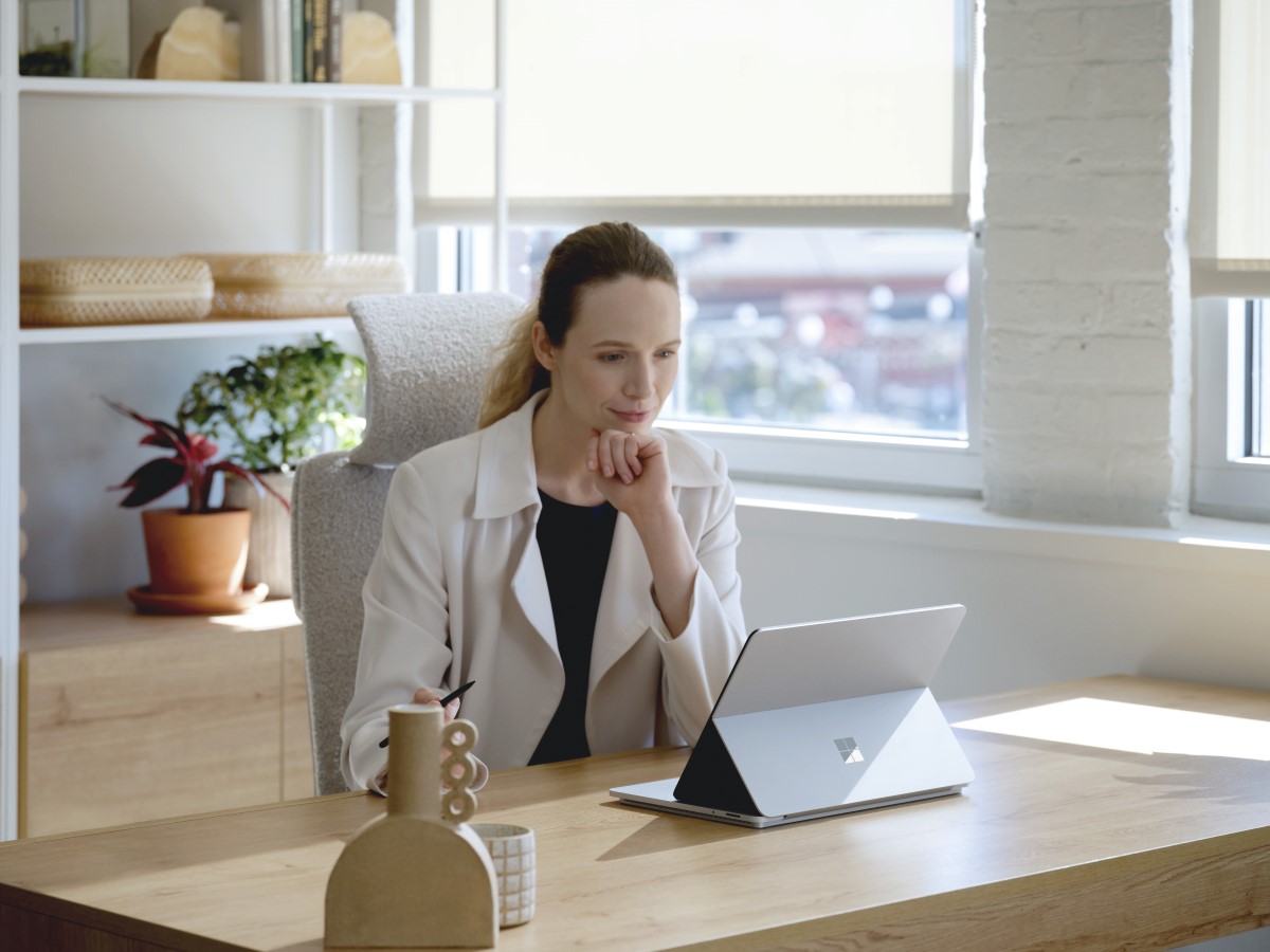 Adult female sitting at a desk, holding a Microsoft Slim Pen 2 while looking onto a platinum Surface Laptop Studio in stage mode