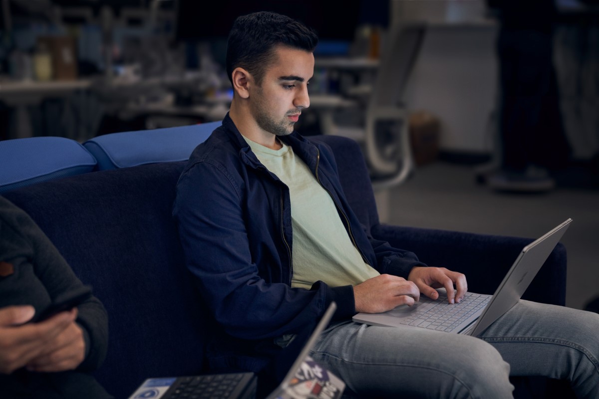 Lightly bearded man working on a laptop while sitting next to another person on a dark blue couch