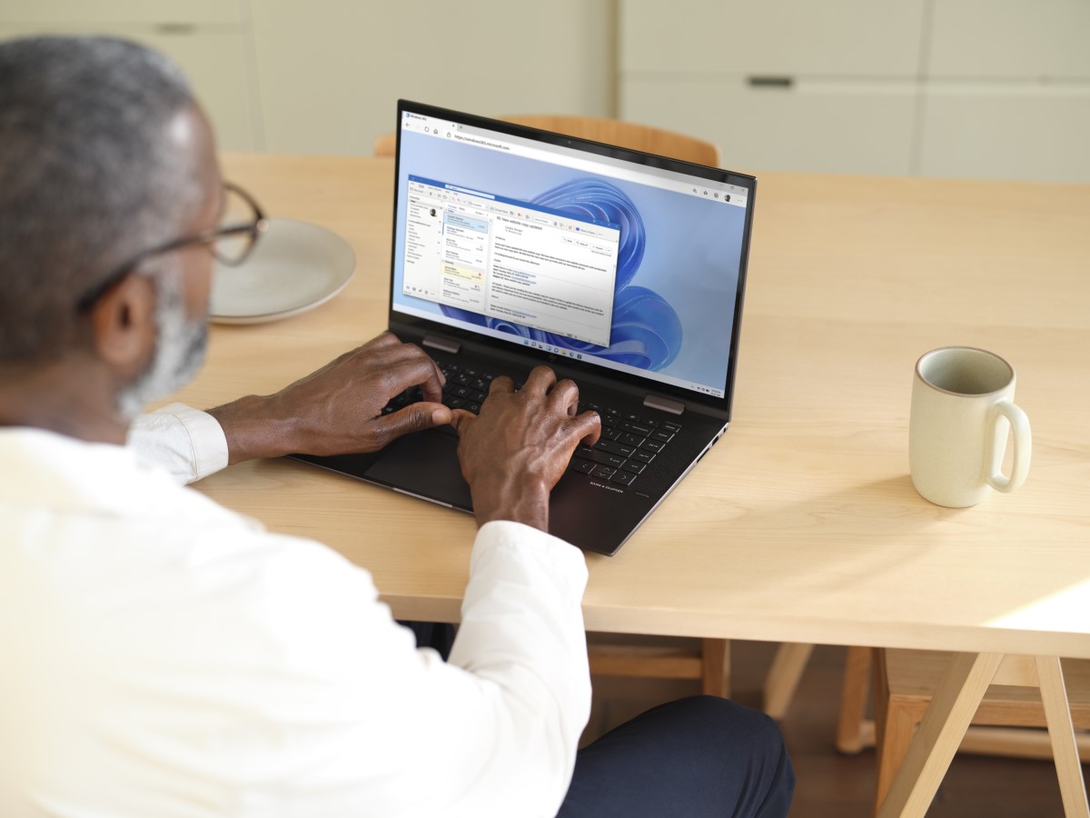 Man sitting at a wooden table typing on a laptop while using Microsoft Teams within Windows 365. Shown on Windows 11