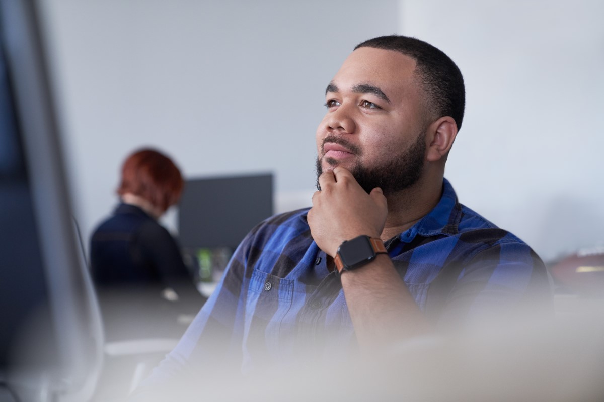 Man with beard sitting behind a computer monitor with his hand to his chin thinking or looking up