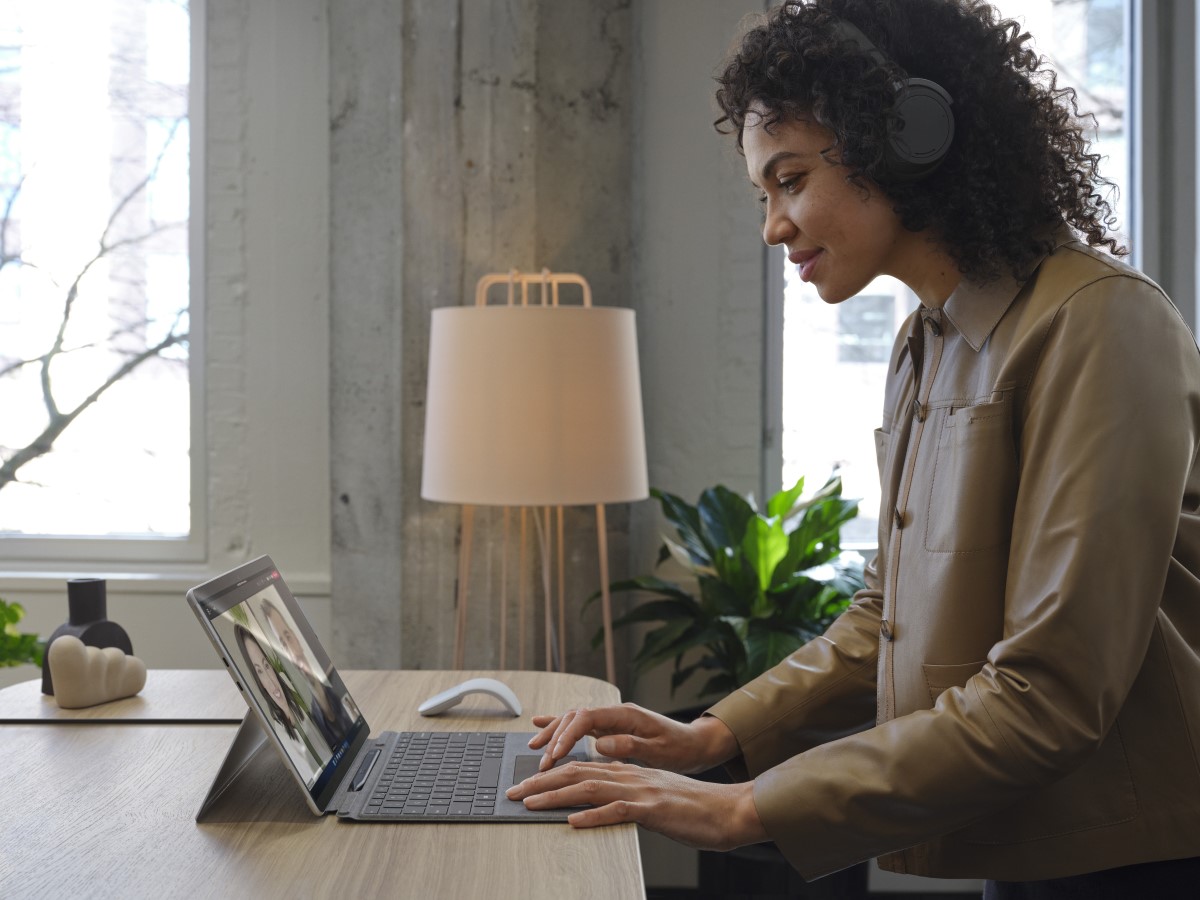 Adult female wearing Surface Headphones 2+ standing at a desk, working on a Surface Pro X in Microsoft Outlook