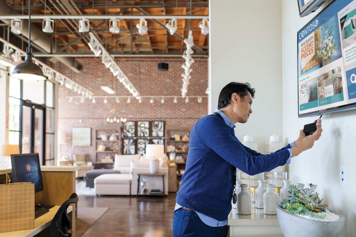 Male employee in commercial retail store plugging Xogo IoT device into wall-mounted monitor. The screen shows product promo ads for home furnishings