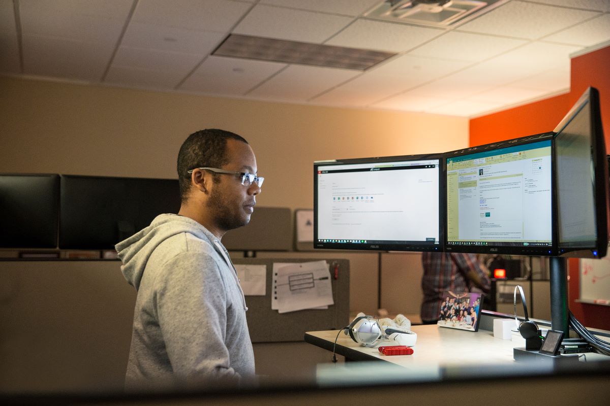 Photo of a male wearing a gray jacket and glasses, standing in front of a computer desk with multiple monitors
