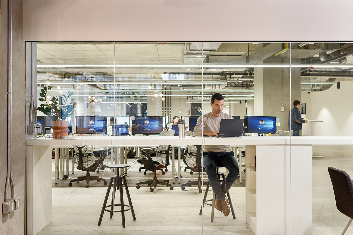 Male office worker seated at high desk in modern office, using Toshiba laptop (screen not visible). Rows of office workstations are in the background, each with desktop computers (some screens partially visible open to Windows 10 log in page)