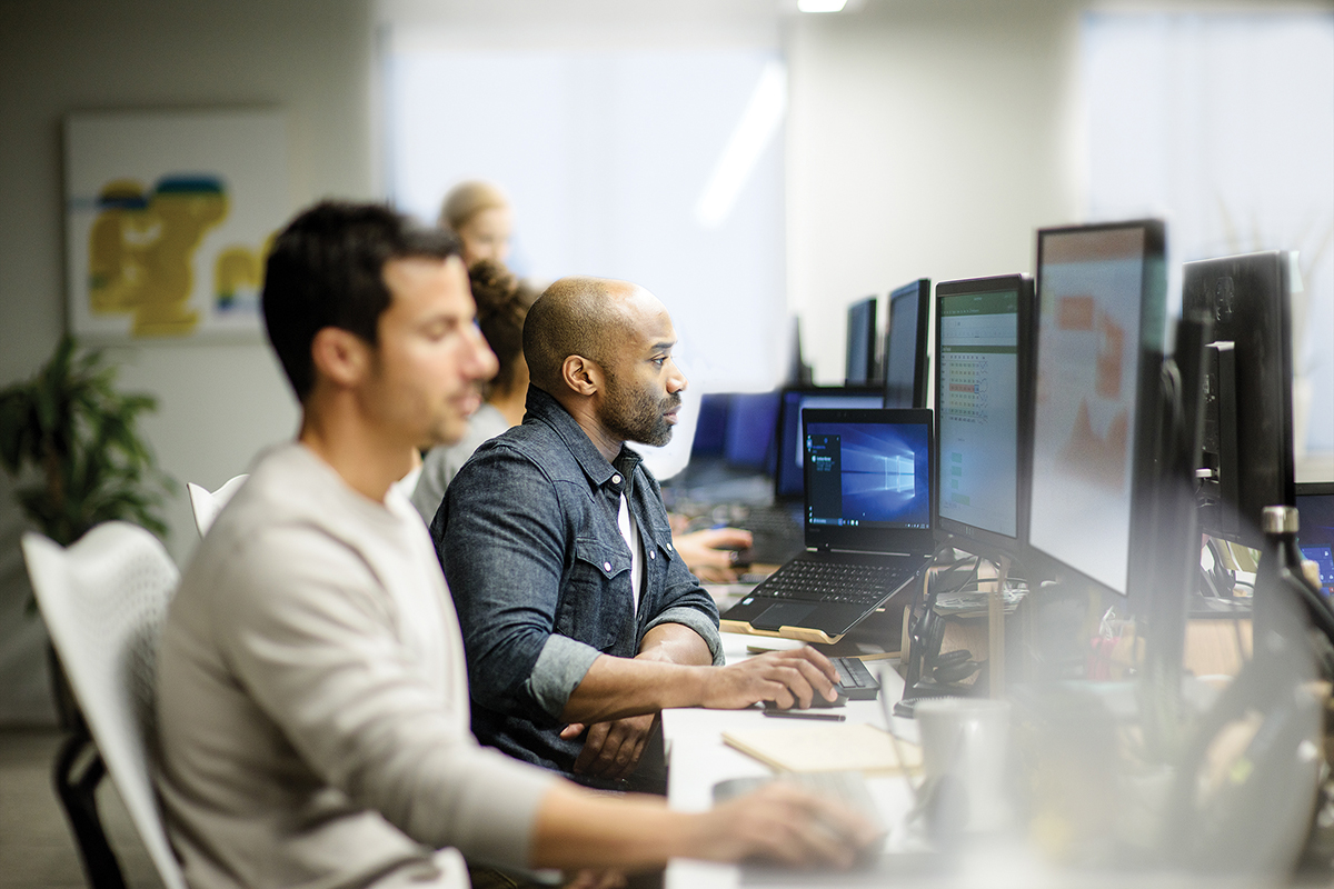 Two male coworkers sitting next to each other along row of office desks, using their own desktop computers (screens show Excel and PowerPoint). An open HP laptop sits on desk to their left, showing the Windows 10 home screen